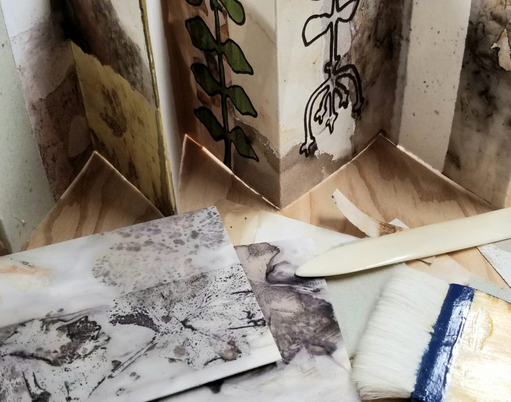 Paper scraps, a glue brush, and a bone folder lying in front of a concertina folded collage.