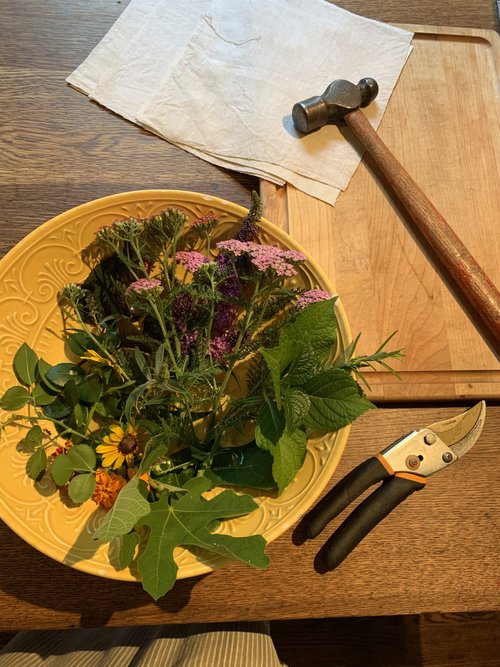 Flowers and leaves in a yellow dish next to white linen, secateurs, and a hammer.